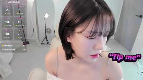 Media: Video of a fair-skinned, short-haired Asian woman in a white dress, looking down, in a modern, minimalist room with white walls and a floor lamp. \"Tip me\" text overlay.