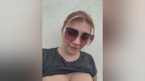 Media: Video of a young Caucasian woman with fair skin, blonde hair, and large pink sunglasses, lifting a grey t-shirt to reveal large, natural breasts. She has a slight smile and is indoors against a textured white wall.