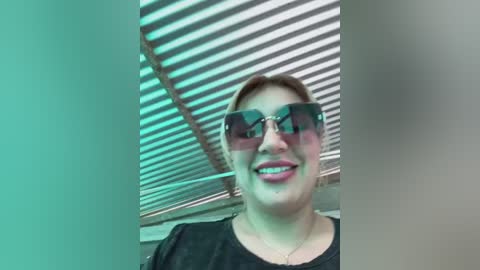 Media: Video of a woman with fair skin, light brown hair, wearing black sunglasses, a black top, and a necklace, standing under a slatted ceiling in a room with green and white lighting.