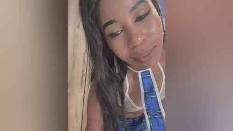 Media: Video of a young Black woman with smooth, medium-brown skin, long dark hair, and a light blue plaid shirt. She has a subtle smile and closed eyes, leaning against a beige wall.