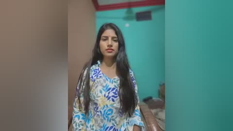 Media: Video of a young woman with long black hair, wearing a colorful floral-patterned blouse, standing in a narrow, brightly lit corridor with teal and peach walls.