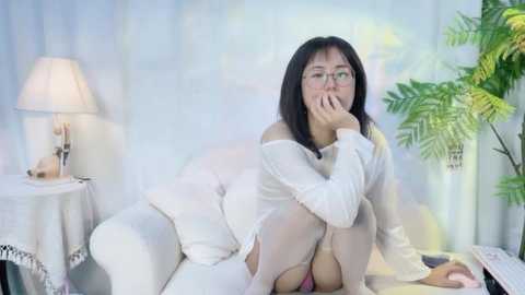 Media: Video of an Asian woman with glasses, sitting on a white couch, wearing a sheer white top, covering her mouth, in a softly lit, white and green-themed room.