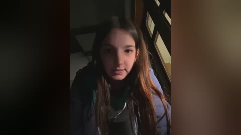 Media: Video of a young girl with long, dark hair, wearing a blue jacket, standing in a dimly lit room with wooden blinds partially closed.