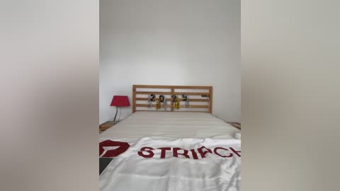 Media: Video of a minimalist, empty bedroom with a wooden bed, red lamp, white wall, and a white bedspread with \"STRAICH\" in red.