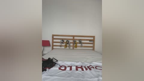 Media: Video of a minimalist, bare room with a wooden bedhead, a white bedsheet, a red chair, a yellow bag, and a white towel with red text, all against a plain white wall.