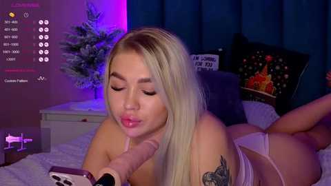 Media: A video shows a blonde woman with light skin, wearing lingerie, lying on a bed, performing oral sex on a large dildo. The room has dark curtains, a Christmas tree, and a \"Cherry Pie\" pillow.