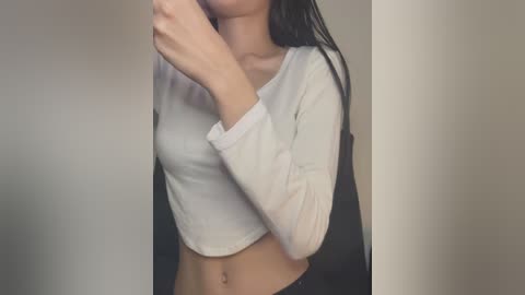 Media: Video of a slim, light-skinned woman with long black hair, wearing a white long-sleeved crop top and black pants, taking a selfie in a plain beige room.