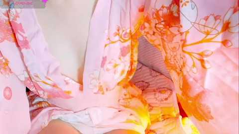 Video of a close-up of a person's chest in a light pink, floral kimono with an open front. The kimono reveals a white bra and light skin tone. The background is blurred, focusing on the garment and undergarment.