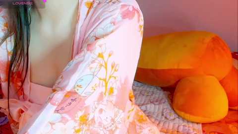 Media: Video of a woman with light skin, wearing a pink floral kimono, lying on a bed with a plush yellow elephant and a striped blanket.