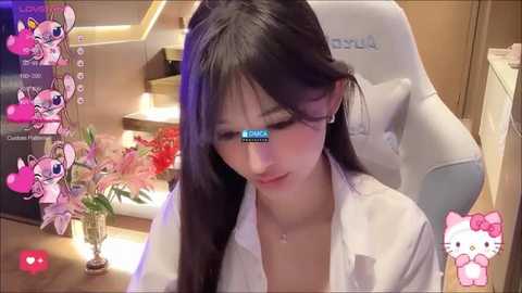 Media: A video of an Asian woman with long black hair, wearing a white blouse, sitting in a white gaming chair. She's surrounded by pink and white anime characters, with a live chat overlay.