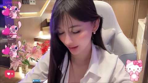 Media: Video of a young Asian woman with long black hair, wearing a white blouse, seated indoors, surrounded by pink Hello Kitty stickers.