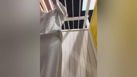 Video of a person in a white robe standing at the top of a spiral staircase with white railings, viewed from below. The background features a yellow wall and a skylight.