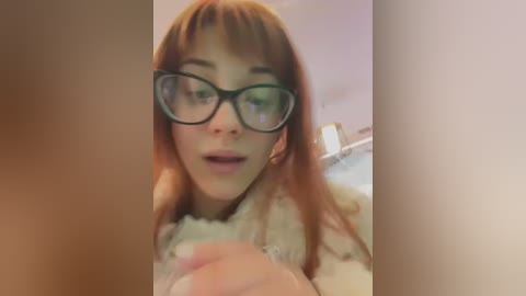 Media: A video of a young woman with long, straight auburn hair, wearing large black-rimmed glasses and a white, fluffy sweater, smiling in a soft-focus bathroom setting.