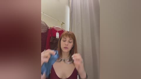 Media: Video of a young woman with light skin and straight brown hair, wearing a burgundy top, adjusting a necklace in a dressing room with red and beige clothes in the background.