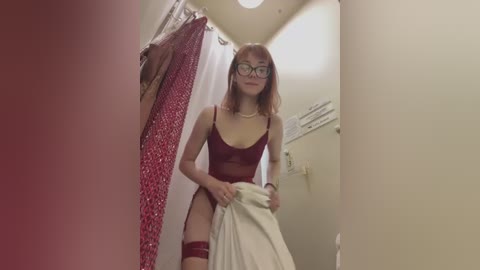 Media: Video of a fair-skinned woman with red hair, glasses, and a maroon lingerie set, standing in a bathroom with red and white curtains, holding a white towel.