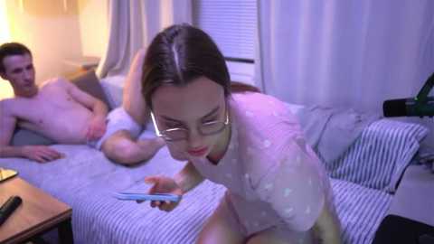 Media: Video of a young woman in glasses, wearing a white blouse, playing with a smartphone while a shirtless man lies on a striped couch in a dimly lit living room.