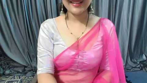 Media: Video of a smiling South Asian woman wearing a white blouse and a sheer pink sari with a subtle floral pattern. She has medium skin tone, gold earrings, and a simple necklace.