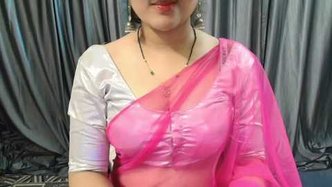 Media: Video of a South Asian woman with light skin, wearing a white and pink sari with a see-through pink dupatta, against a gray, draped fabric backdrop.