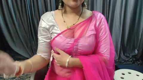 Media: Video of a woman in a semi-sheer pink and white sari, with a black dupatta, against gray curtains. She wears large gold earrings and a necklace.