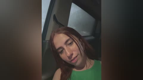 Media: A video of a young Caucasian woman with long, straight auburn hair, wearing a green tank top, sitting in a car. Her eyes are closed, and she appears relaxed. The car's interior is dimly lit, with a gray seat visible in the background.