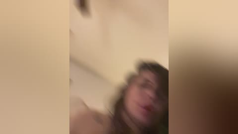 Media: Blurry video of a nude woman with brown hair, partially visible from the waist up, against a beige background. Her expression is difficult to discern due to the lack of focus.