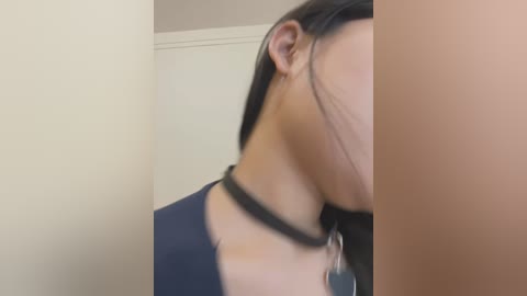 Media: A video showing a side profile of a woman with medium-length black hair, wearing a black choker with a pendant, and a navy blue top. The background features a beige wall with a white trim.