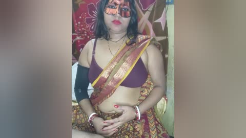 Media: Video of a woman in traditional Indian attire, wearing a maroon bra, a red sari, and a golden sash. She has a black eye patch and a cast on her left arm. The background features floral tapestries.