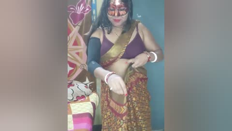 Media: Video of a South Asian woman in a traditional sari, with an ornate golden shoulder drape, and a colorful, patterned skirt. She has a gold mask and a black arm brace, standing against a vibrant, floral-patterned wall.