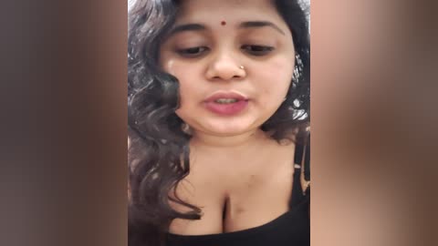 Media: A video of a young South Asian woman with medium skin tone, curly black hair, and a small red bindi. She wears a black top, revealing cleavage. The background is blurred.