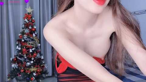 Media: A video of a fair-skinned, long-haired woman with red lipstick, wearing a strapless dress with a red bow, in a room with a decorated Christmas tree and grey curtains.