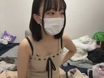 Media: Video of an Asian woman with straight black hair and bangs, wearing a beige ribbed tank top, white mask, and black bra straps, sitting on a bed surrounded by disheveled clothes and stuffed animals.