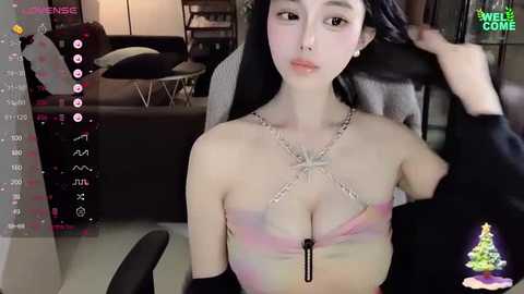 Media: A video of a young, fair-skinned Asian woman with long black hair, wearing a revealing, pastel-colored halter top and a silver necklace, seated in a dimly lit room with a Christmas tree and a gaming setup in the background.