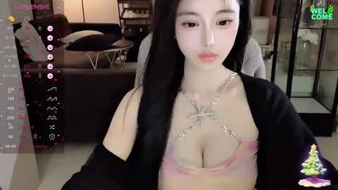 Media: Video of an East Asian woman with long black hair, fair skin, and wearing a revealing pink bra with a black blazer. Background shows a modern living room with a TV screen displaying social media features.