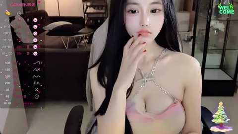 Media: Video of an East Asian woman with long black hair, wearing a pink bra with silver chains, sitting on a gaming chair in a modern living room.