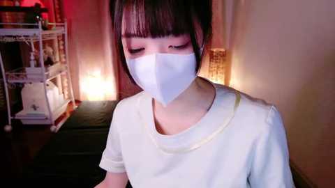 Media: Video of a young Asian woman with straight black hair, wearing a white mask and T-shirt, standing in a dimly lit room with a red glow and a white metal shelving unit in the background.