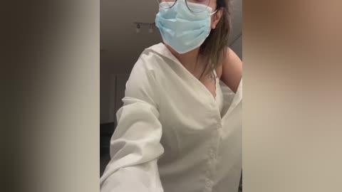 Media: Video of a woman wearing a white button-up shirt, blue mask, and glasses, partially revealing a black bra strap, standing in a dimly lit, white-walled hallway.