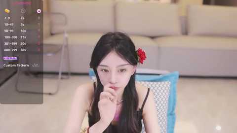 Media: Video of an East Asian woman with long black hair, red flower accessory, and fair skin, sitting on a blue and white patterned rug in a modern living room with beige couches.