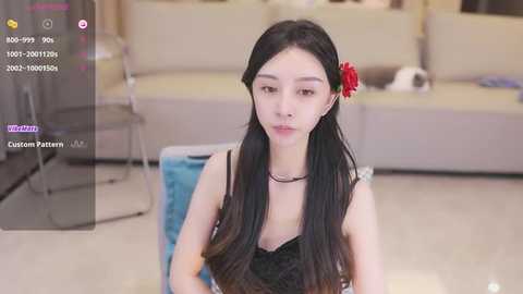 Media: Video of an Asian woman with long black hair, wearing a black lace top and red flower, sitting on a blue cushion in a modern living room.