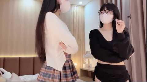 Media: A video shows two East Asian women in a modern bedroom. One wears a white sweater and plaid skirt, the other a black crop top and skirt. They both have long black hair and are wearing face masks.