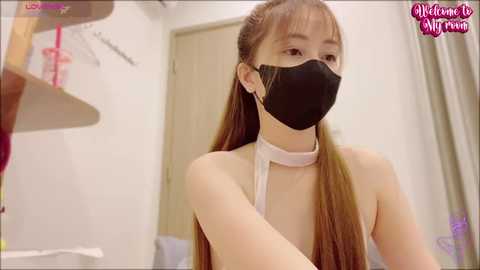 Media: Video of an East Asian woman with long brown hair, wearing a black face mask and white apron, cleaning a white bathroom sink.