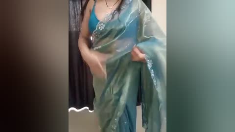 Media: Video of a woman in a teal, semi-transparent saree with intricate embroidery, posing indoors. She has medium skin tone, long dark hair, and is wearing a matching blouse. The background features a dark curtain and light-colored walls.