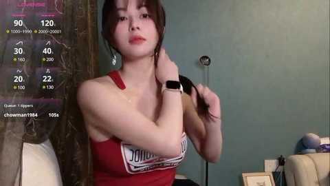 Media: A video of a young Asian woman with straight black hair and fair skin, wearing a red tank top, sitting in a teal room, holding a microphone.