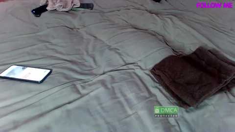 Media: A video of a beige quilted bedspread with a brown blanket folded on one side. A smartphone and a black hairbrush lie on the bed, with a green and purple logo at the bottom right.