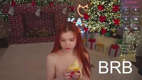 Media: Video of a nude, fair-skinned woman with long red hair, holding a sandwich, in a festive, decorated living room with Christmas lights and a fireplace.