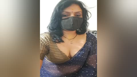 Media: Video of a South Asian woman with medium skin tone, wearing a black mask, dark blue saree, and a beige top, partially visible cleavage, in a plain indoor background.