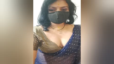 Media: Video of a South Asian woman with medium-dark skin, wearing a black mask, a gold blouse with a deep V-neck, and a sheer blue sari with white polka dots. She has shoulder-length black hair and a bindi on her forehead.