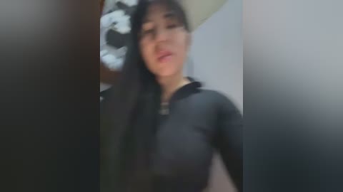 Media: Blurry video of an East Asian woman with long black hair, wearing a black top, standing indoors near a white wall with a white flower on it.