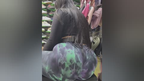 Media: Video of a curvy, dark-skinned woman with long, wavy hair, wearing a black crop top and tight, green and purple tie-dye leggings. She sits on a bench in a crowded, dimly lit indoor setting.