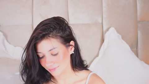 Media: Video of a young woman with long, dark hair, wearing a white tank top, smiling gently, seated on a bed with white pillows and beige upholstered headboard in a softly lit room.