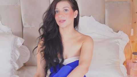 Media: Video of a fair-skinned woman with long, wavy black hair, wearing a blue strapless dress, sitting on a bed with white pillows and beige headboard.